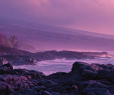 紫色の光に包まれたハワイ島の海岸の風景。溶岩の黒い岩場と静かな波が広がり、夜明け前の淡い光が大地を照らす静寂の景色を描いたイメージ。